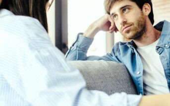 A young man with a beard sits on a couch, resting his head on his hand and looking thoughtfully at a woman sitting across from him. Both appear to be engaged in a serious conversation.