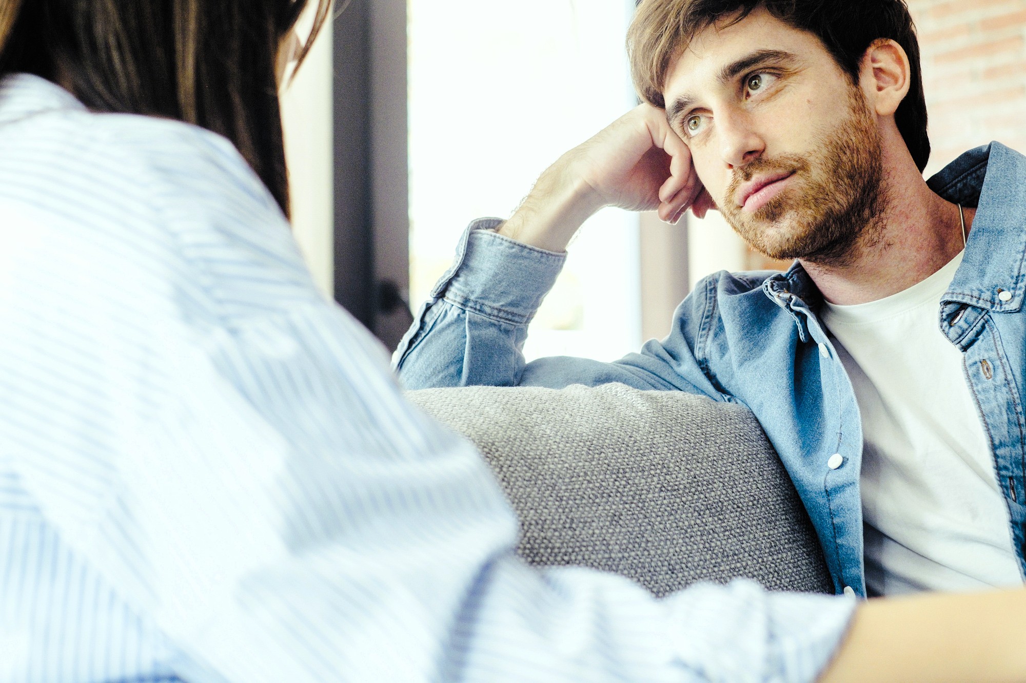 A young man with a beard sits on a couch, resting his head on his hand and looking thoughtfully at a woman sitting across from him. Both appear to be engaged in a serious conversation.