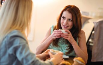 Two women sit at a table in a café, holding cups and engaged in conversation. One woman with long red hair smiles while listening, while the other, blonde, is seen from behind. Muffins are on the table.