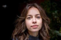 A young woman with wavy brown hair, clear skin, and blue eyes looks directly at the camera with a neutral expression. She is wearing a dark sweater and standing in front of a blurred, dark background with greenery.