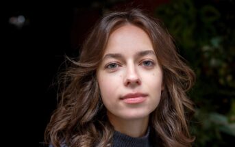 A young woman with wavy brown hair, clear skin, and blue eyes looks directly at the camera with a neutral expression. She is wearing a dark sweater and standing in front of a blurred, dark background with greenery.