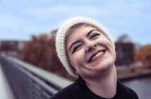 A person wearing a white knit beanie and dark clothing smiles broadly outdoors, showing their teeth. They have short hair, a nose ring, bold eyeliner, and are standing on a bridge with blurred trees and sky in the background.