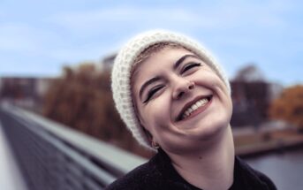 A person wearing a white knit beanie and dark clothing smiles broadly outdoors, showing their teeth. They have short hair, a nose ring, bold eyeliner, and are standing on a bridge with blurred trees and sky in the background.