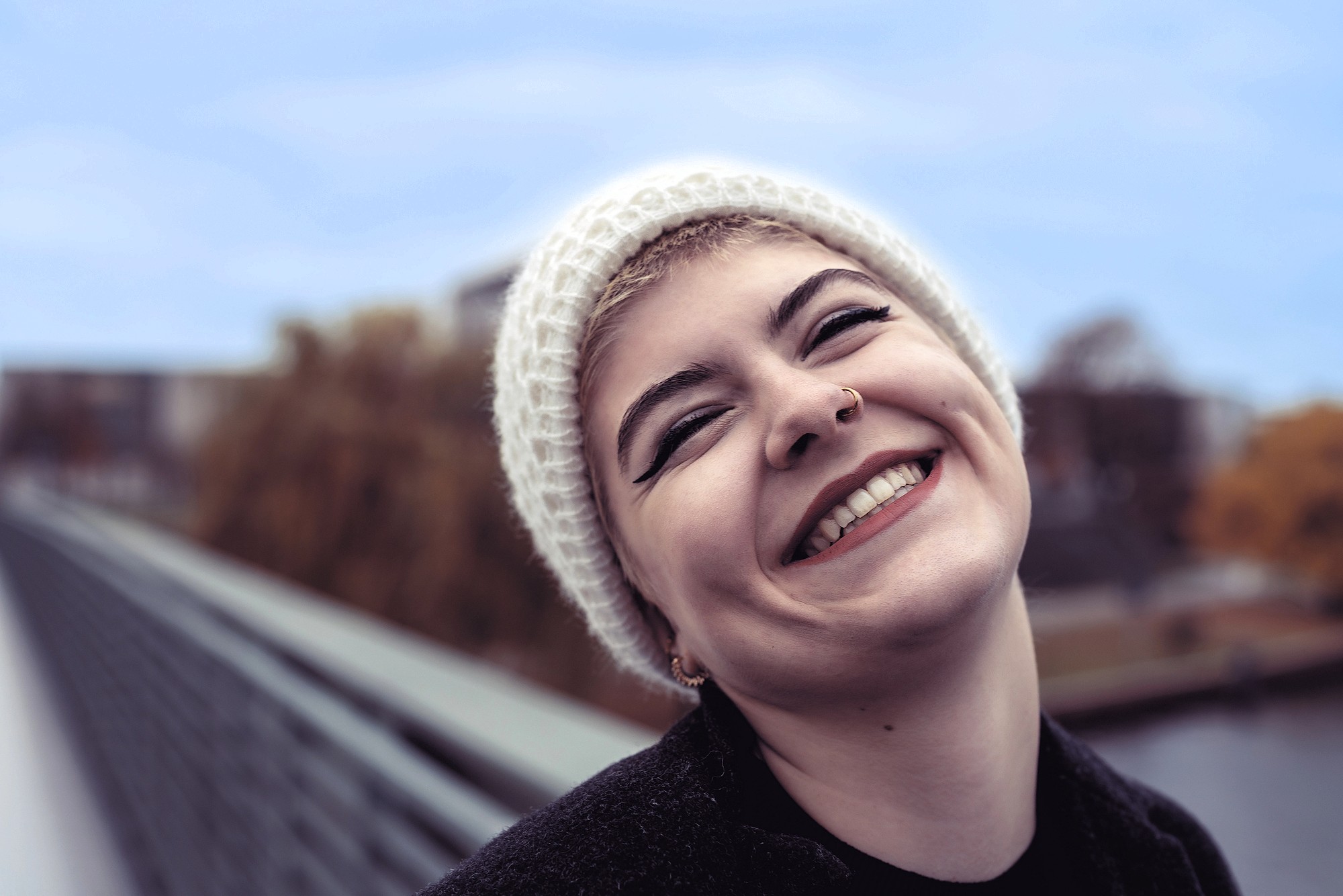 A person wearing a white knit beanie and dark clothing smiles broadly outdoors, showing their teeth. They have short hair, a nose ring, bold eyeliner, and are standing on a bridge with blurred trees and sky in the background.