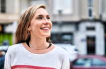 A woman with long hair smiles and looks up while standing outdoors. She is wearing a white sweater with pink and red stripes, and the background shows a blurred city street scene.
