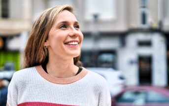 A woman with long hair smiles and looks up while standing outdoors. She is wearing a white sweater with pink and red stripes, and the background shows a blurred city street scene.