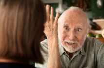 An elderly man with white hair and a beard gestures expressively with his hand while talking to a woman whose back is to the camera. The man appears engaged and animated in conversation.