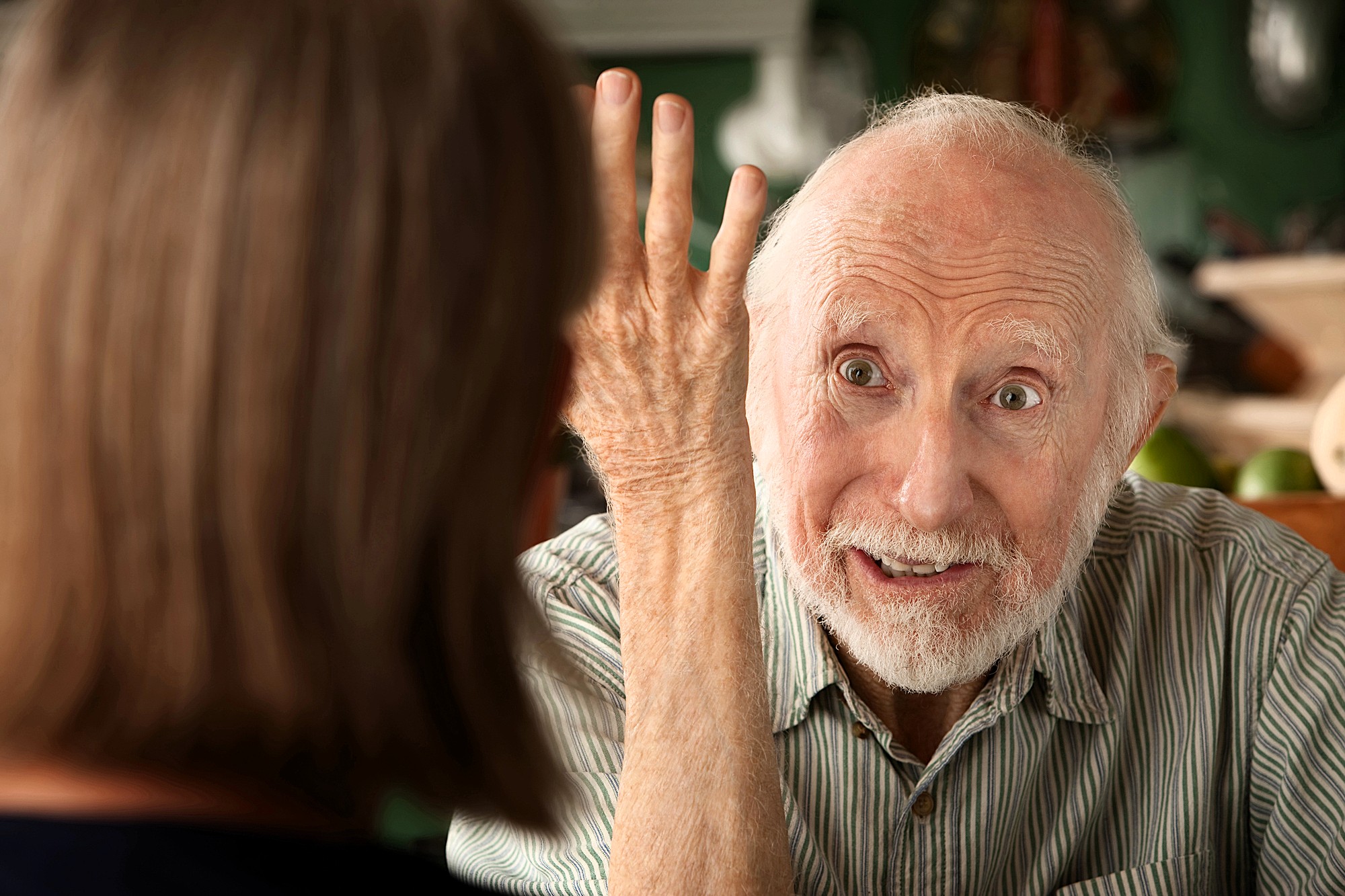 An elderly man with white hair and a beard gestures expressively with his hand while talking to a woman whose back is to the camera. The man appears engaged and animated in conversation.