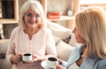 Two women, one older with gray hair and glasses and one younger with blonde hair, sit on a couch holding cups of coffee, smiling and chatting in a cozy living room.