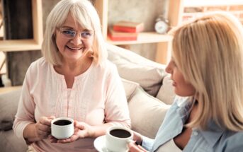 Two women, one older with gray hair and glasses and one younger with blonde hair, sit on a couch holding cups of coffee, smiling and chatting in a cozy living room.