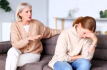 An older woman sits on a couch, talking and offering comfort to a younger woman who appears upset, resting her head on her hand and looking down. Both are dressed in neutral sweaters in a bright, homey room.