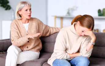 An older woman sits on a couch, talking and offering comfort to a younger woman who appears upset, resting her head on her hand and looking down. Both are dressed in neutral sweaters in a bright, homey room.