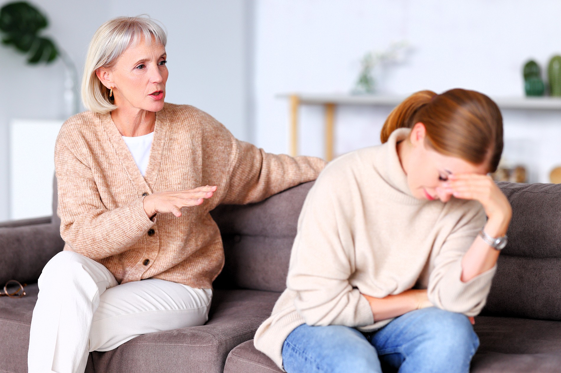An older woman sits on a couch, talking and offering comfort to a younger woman who appears upset, resting her head on her hand and looking down. Both are dressed in neutral sweaters in a bright, homey room.