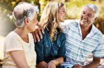 Three people sit outdoors, smiling and talking. An older man and woman sit on either side of a younger woman. The man has his arm around the younger woman. The background is sunny and filled with greenery.