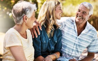 Three people sit outdoors, smiling and talking. An older man and woman sit on either side of a younger woman. The man has his arm around the younger woman. The background is sunny and filled with greenery.