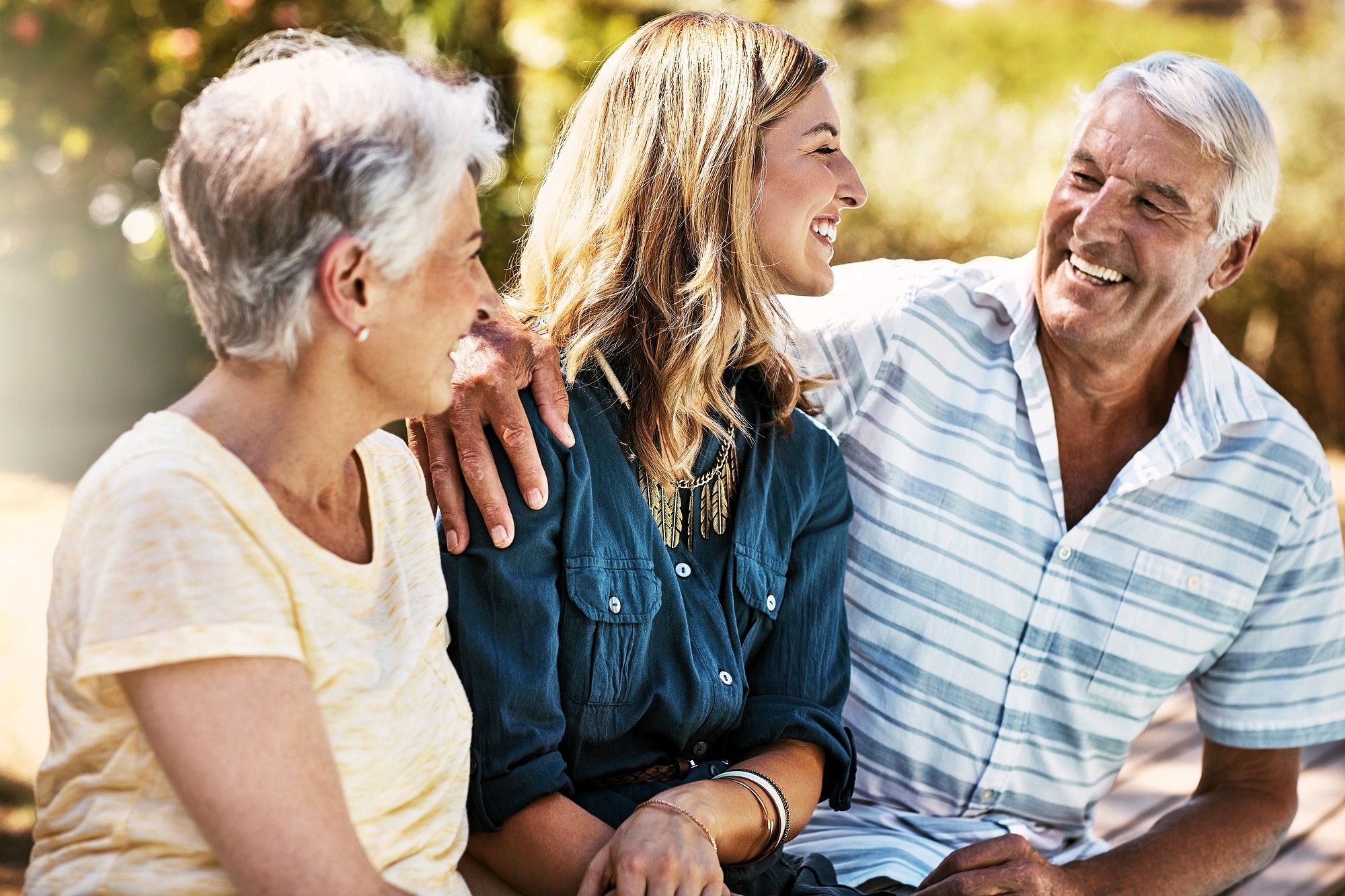 Three people sit outdoors, smiling and talking. An older man and woman sit on either side of a younger woman. The man has his arm around the younger woman. The background is sunny and filled with greenery.