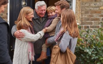 A smiling older couple greets a family, including a young child, at the front door of a brick house. They embrace warmly, suggesting a happy reunion. Everyone is dressed in cozy, autumn clothing.