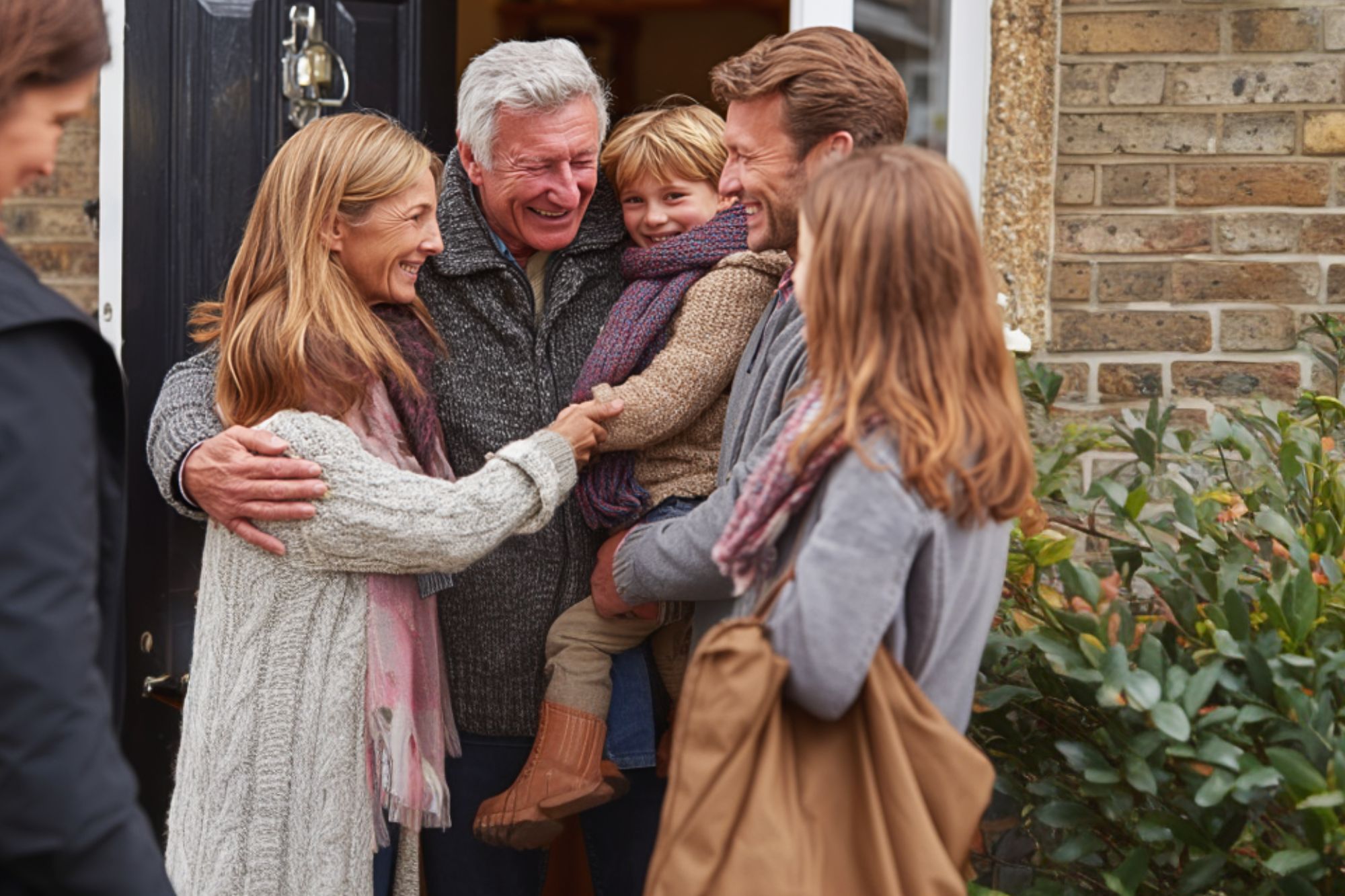 A smiling older couple greets a family, including a young child, at the front door of a brick house. They embrace warmly, suggesting a happy reunion. Everyone is dressed in cozy, autumn clothing.
