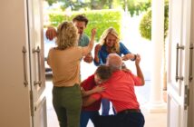 A joyful family reunion at a house entrance, with adults greeting each other warmly and a young boy hugging an older man. Sunlight shines in, and greenery is visible outside.