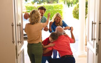 A joyful family reunion at a house entrance, with adults greeting each other warmly and a young boy hugging an older man. Sunlight shines in, and greenery is visible outside.