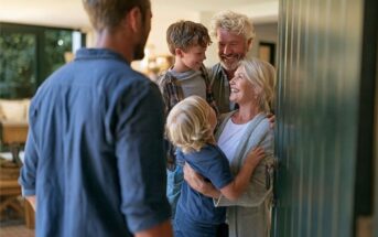 A man greets two smiling grandparents and two children at the door of a home. The grandparents are hugging the kids and all appear happy, suggesting a family reunion or visit.