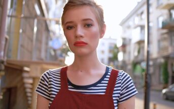 A young person with short hair wears a striped shirt under a red overall, standing outside on a city street with a neutral expression. The background shows blurred buildings and bright sunlight.
