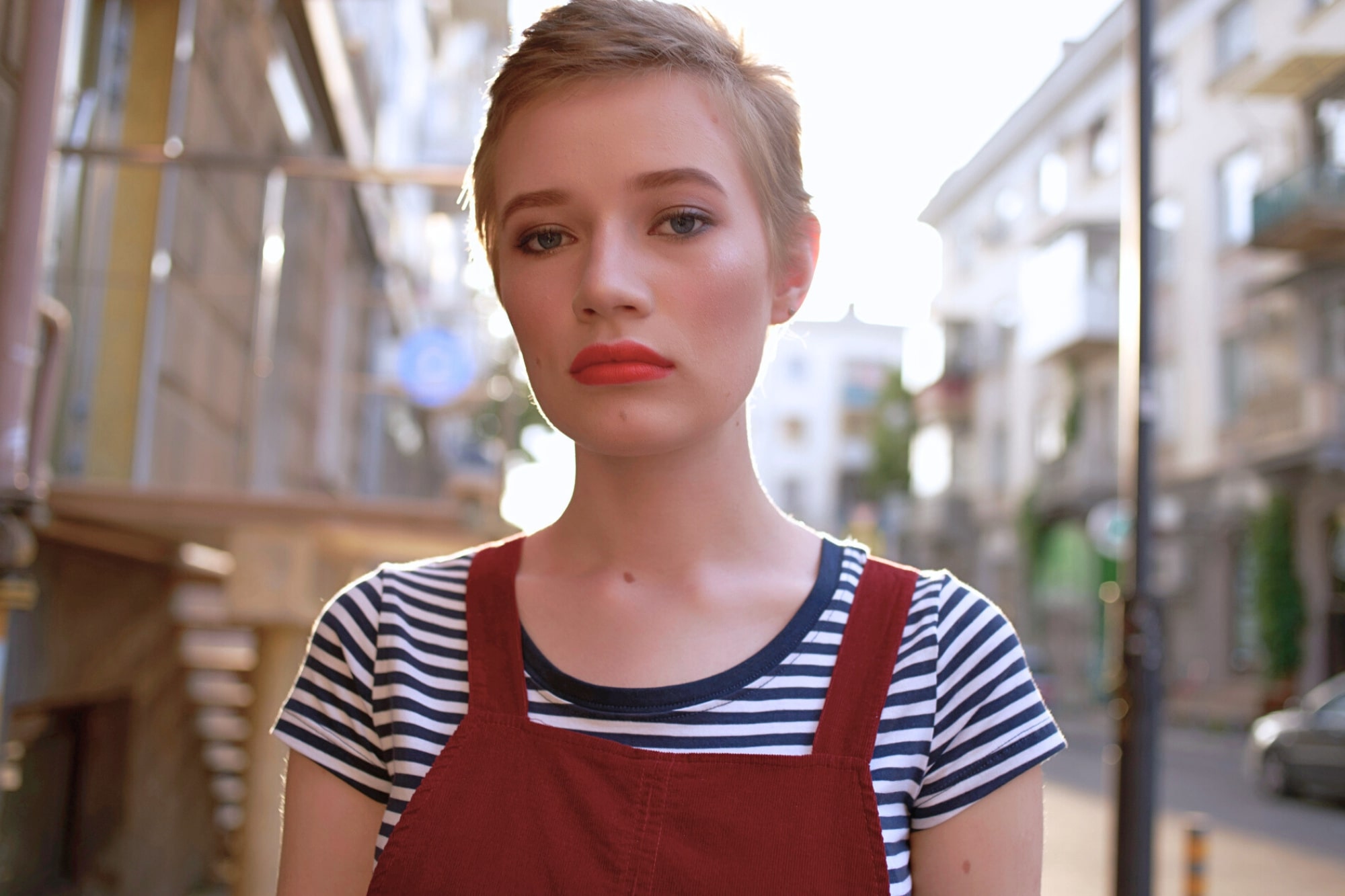 A young person with short hair wears a striped shirt under a red overall, standing outside on a city street with a neutral expression. The background shows blurred buildings and bright sunlight.