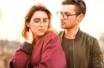 A young woman in a red jacket and glasses looks down while a young man in a green jacket and glasses stands close beside her, gazing at her with a soft expression. They are outdoors in natural light.