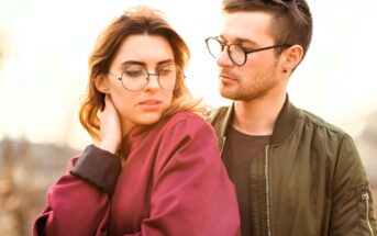 A young woman in a red jacket and glasses looks down while a young man in a green jacket and glasses stands close beside her, gazing at her with a soft expression. They are outdoors in natural light.