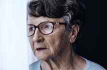 An elderly woman with short gray hair and glasses looks thoughtfully out a window, her face partially illuminated by soft light, with a neutral expression.
