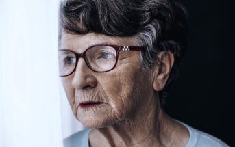 An elderly woman with short gray hair and glasses looks thoughtfully out a window, her face partially illuminated by soft light, with a neutral expression.