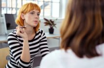 A woman with short red hair in a black-and-white striped shirt holds a pen and listens attentively to another person during a conversation in a bright office setting.