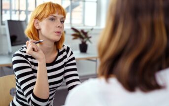 A woman with short red hair in a black-and-white striped shirt holds a pen and listens attentively to another person during a conversation in a bright office setting.