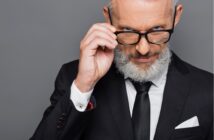 A confident older man with gray hair and a beard, wearing a black suit and tie, adjusts his glasses while looking intently at the camera against a gray background.