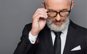 A confident older man with gray hair and a beard, wearing a black suit and tie, adjusts his glasses while looking intently at the camera against a gray background.