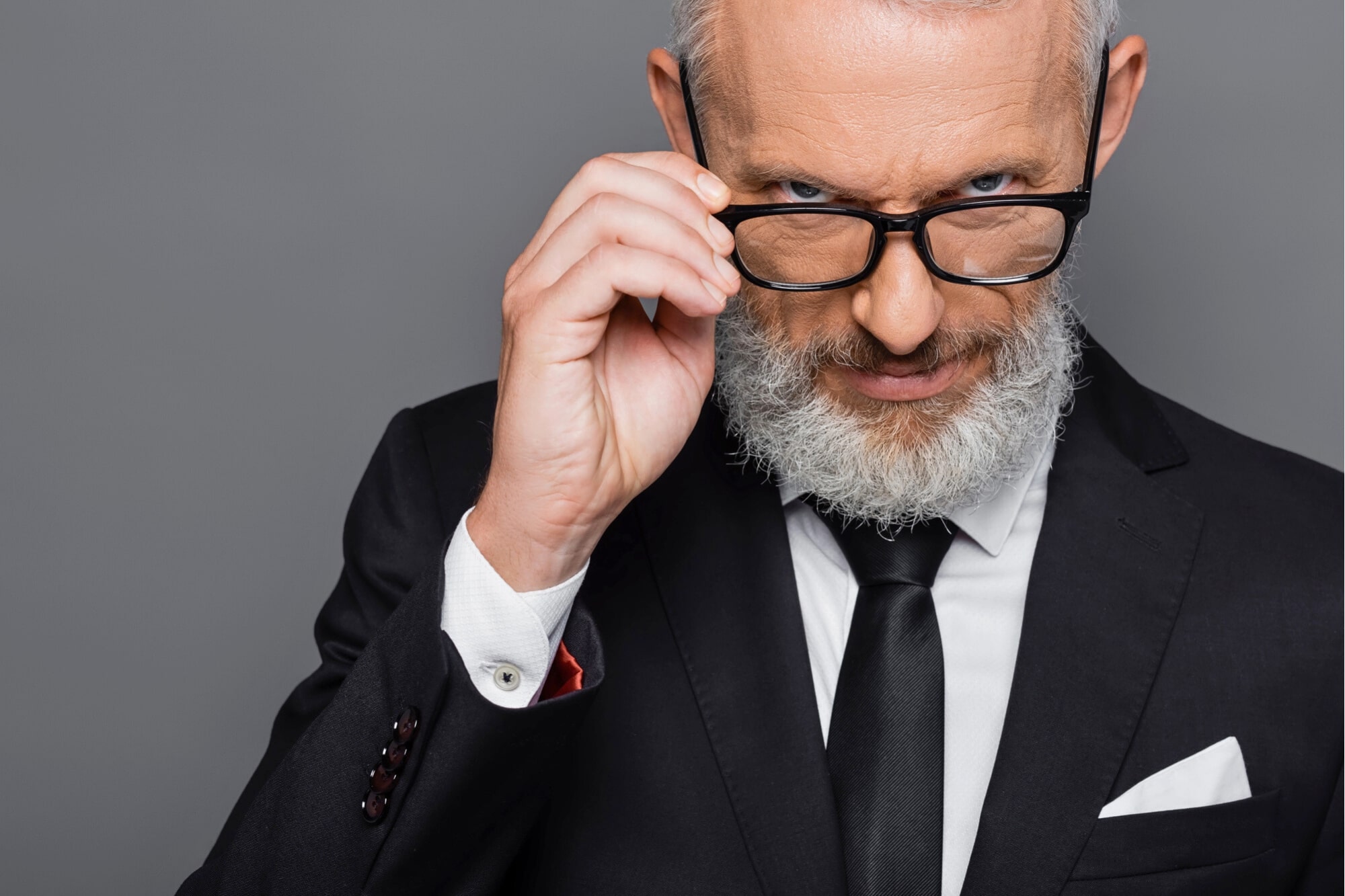 A confident older man with gray hair and a beard, wearing a black suit and tie, adjusts his glasses while looking intently at the camera against a gray background.