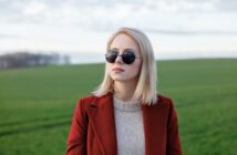 A woman with straight blonde hair wearing round sunglasses, a beige sweater, and a red coat stands outdoors in a green field with a cloudy sky in the background.