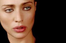 A close-up of a woman with pale skin, blue eyes, and pink lips looking slightly to the side against a black background.