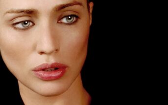 A close-up of a woman with pale skin, blue eyes, and pink lips looking slightly to the side against a black background.