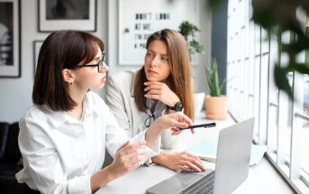 Two women sit at a desk by a window with a laptop, having a focused discussion. One woman gestures with a pen while the other listens attentively. The setting is modern and bright, with plants and framed art in the background.