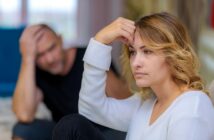 A woman with blonde hair looks upset while sitting indoors, resting her forehead on her hand. In the blurred background, a man in a black shirt appears distressed, sitting with his hand on his head.
