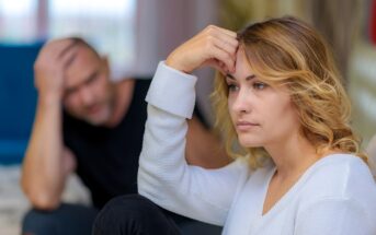 A woman with blonde hair looks upset while sitting indoors, resting her forehead on her hand. In the blurred background, a man in a black shirt appears distressed, sitting with his hand on his head.