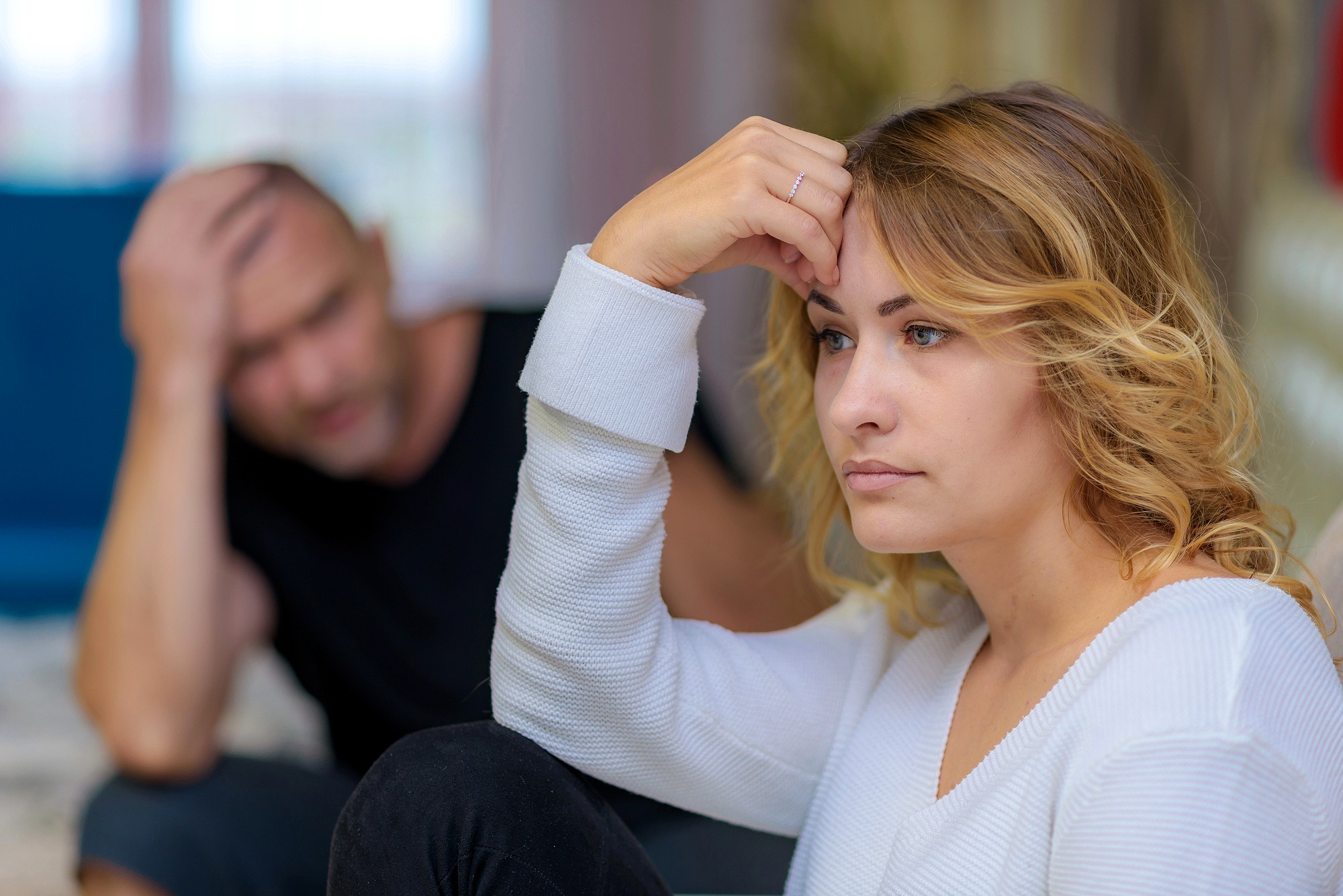A woman with blonde hair looks upset while sitting indoors, resting her forehead on her hand. In the blurred background, a man in a black shirt appears distressed, sitting with his hand on his head.