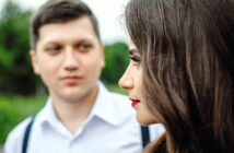 A close-up of a woman in profile with long brown hair and red lipstick, standing outdoors, while a man in a white shirt with suspenders looks at her in the blurred background.