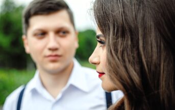 A close-up of a woman in profile with long brown hair and red lipstick, standing outdoors, while a man in a white shirt with suspenders looks at her in the blurred background.