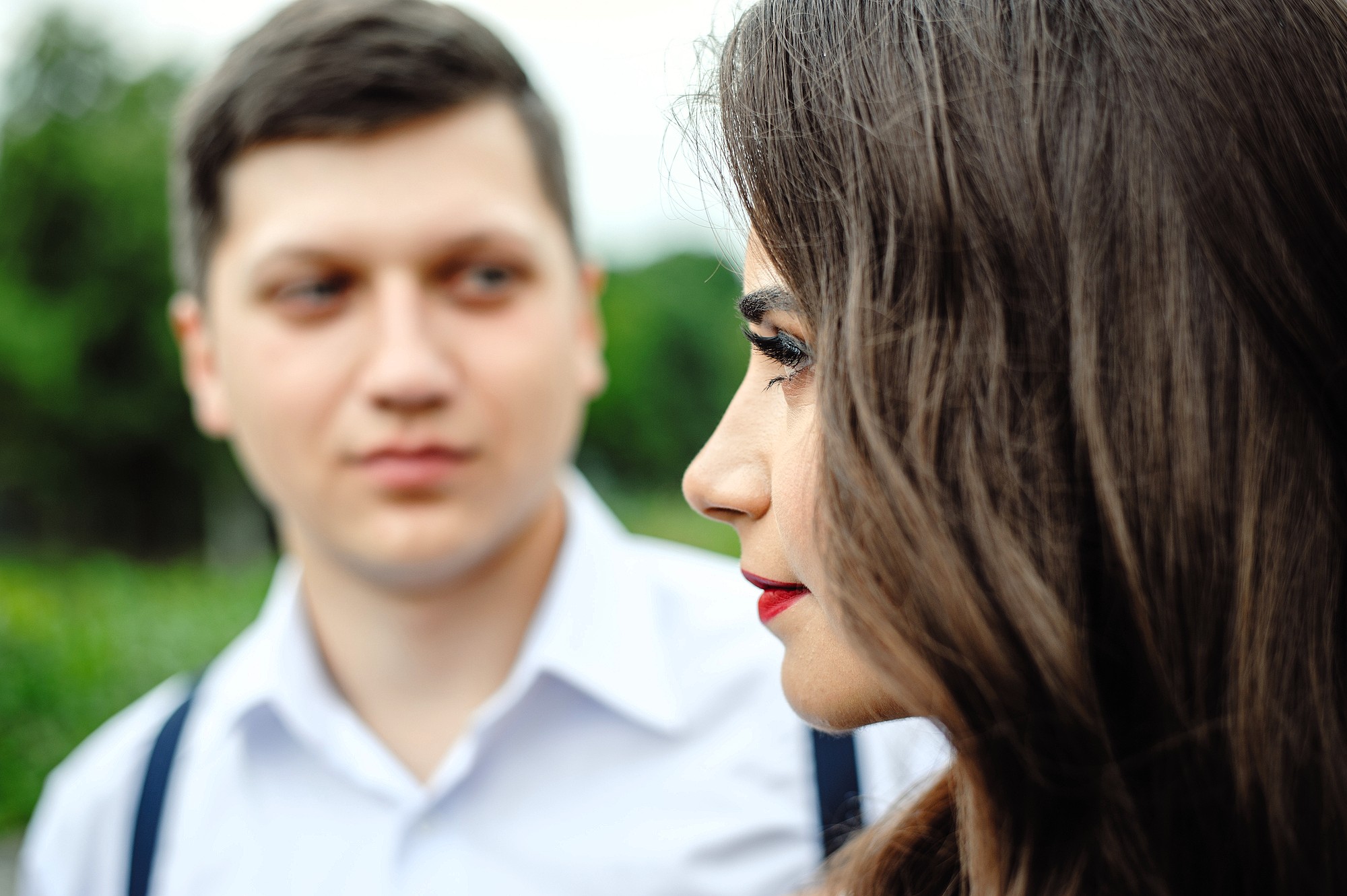 A close-up of a woman in profile with long brown hair and red lipstick, standing outdoors, while a man in a white shirt with suspenders looks at her in the blurred background.