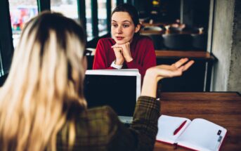 Two women sit across from each other at a table in a cafe. One faces the camera, listening with her chin on her hands; the other gestures while speaking. A laptop and open notebook lie on the table.