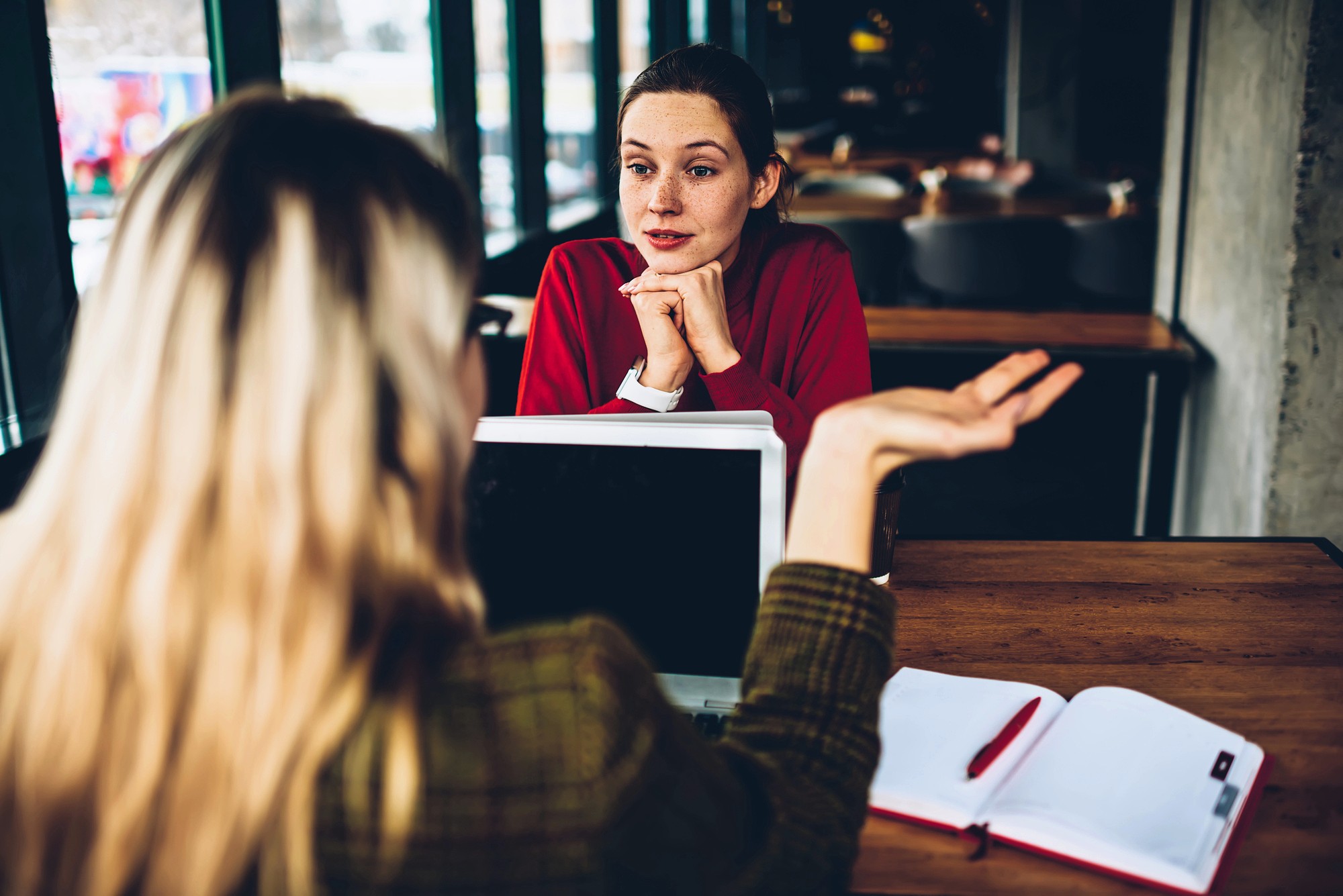 Two women sit across from each other at a table in a cafe. One faces the camera, listening with her chin on her hands; the other gestures while speaking. A laptop and open notebook lie on the table.