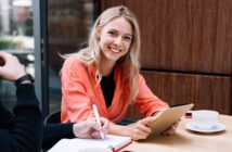 A smiling woman with long blonde hair wearing an orange shirt sits at a table holding a clipboard. Another person is writing in a notebook. A cup of coffee is on the table beside them.