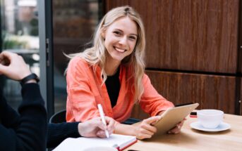 A smiling woman with long blonde hair wearing an orange shirt sits at a table holding a clipboard. Another person is writing in a notebook. A cup of coffee is on the table beside them.